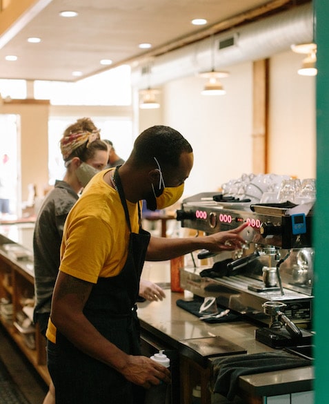 El wears an orange mask and shirt, making an espresso behind the bar at Blueprint Coffee Delmar