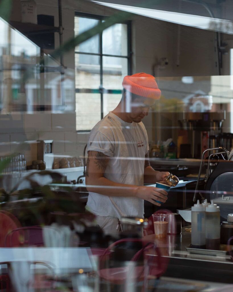 britton watson cafe Britton steaming milk at the espresso machine at Watson, viewed from outside looking in through the window