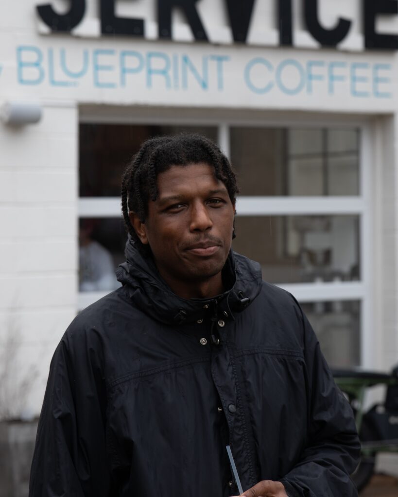 Micah stands in front of our Watson café holding an iced drink, smiling at the camera