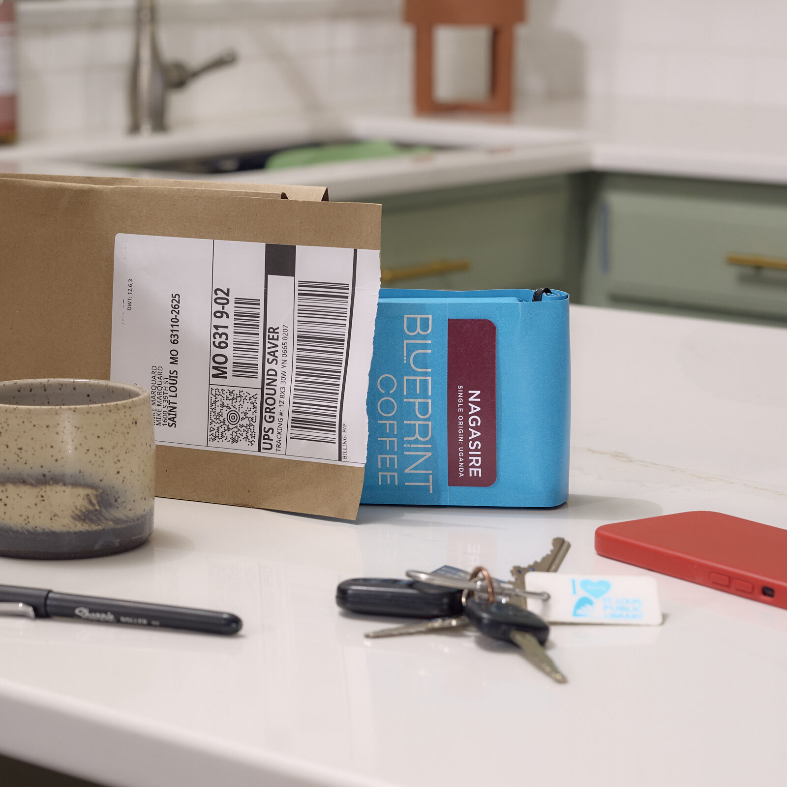 An open subscription mailing envelope lies on a home kitchen counter, with a blue 10 oz bag of Blueprint Coffee beans halfway removed. A set of keys, cell phone, coffee mug, and ballpoint pen sit nearby.