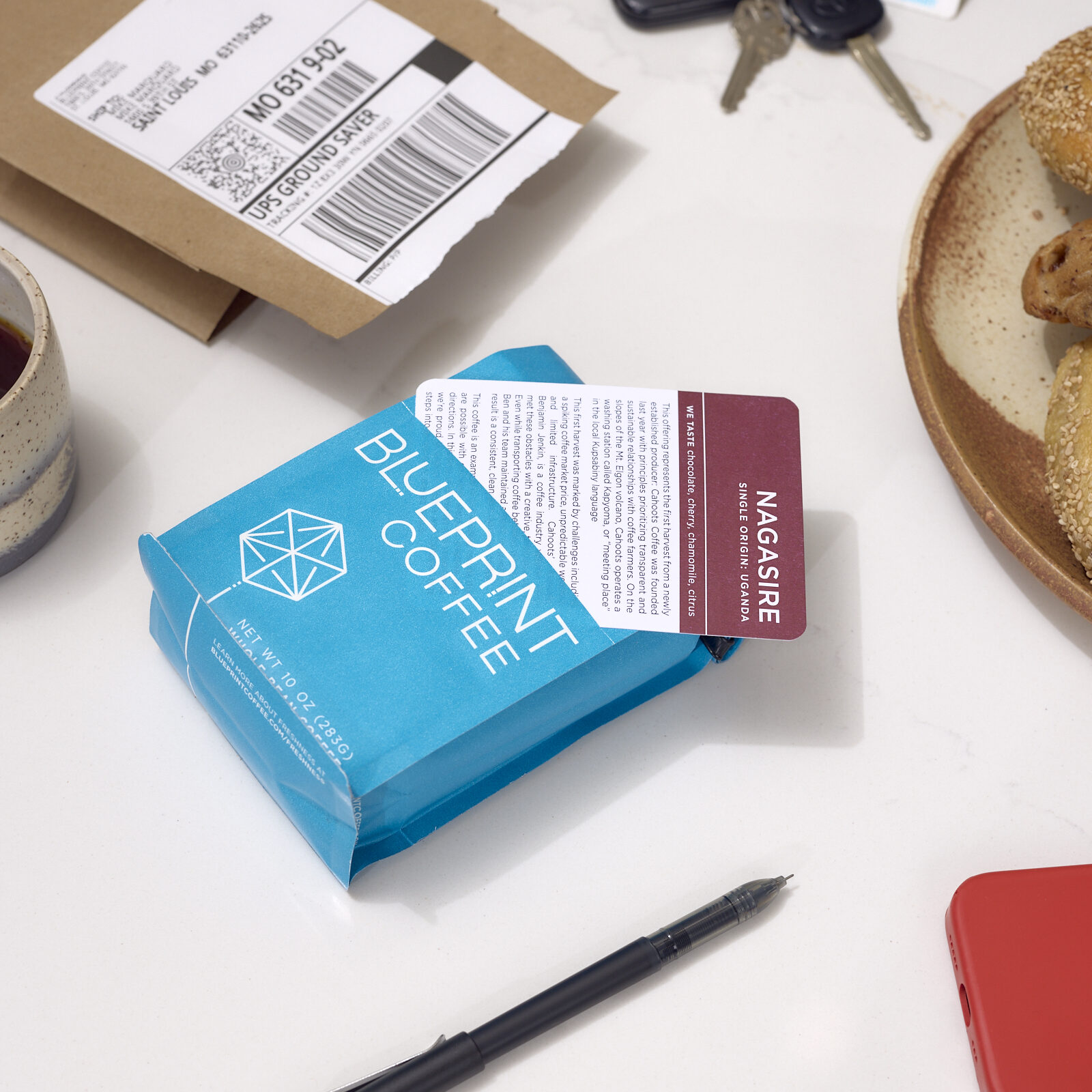 A blue 10 oz bag of Blueprint Coffee beans lies on a home kitchen counter, with an open mailing envelope, ballpoint pen, and set of keys nearby. The removable coffee info card is partially removed from the bag's front pocket, showing the coffee's sourcing notes printed on the card.