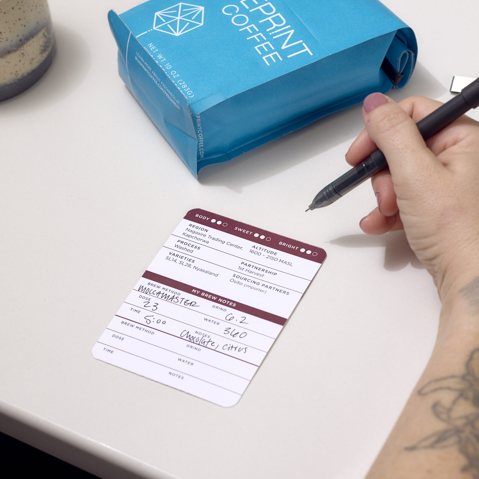 A person writes tasting notes on the removable info card that's been pulled from the front pocket of a blue 10 oz bag of Blueprint Coffee beans, which sits nearby