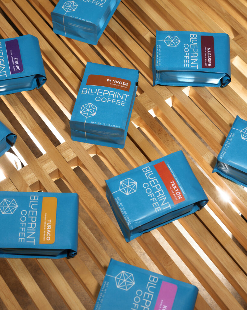 Bright blue coffee bags with the Blueprint Coffee logo are arranged on a wooden surface. Each bag includes a removable card printed with various bright colors like orange, yellow, purple, and brown.