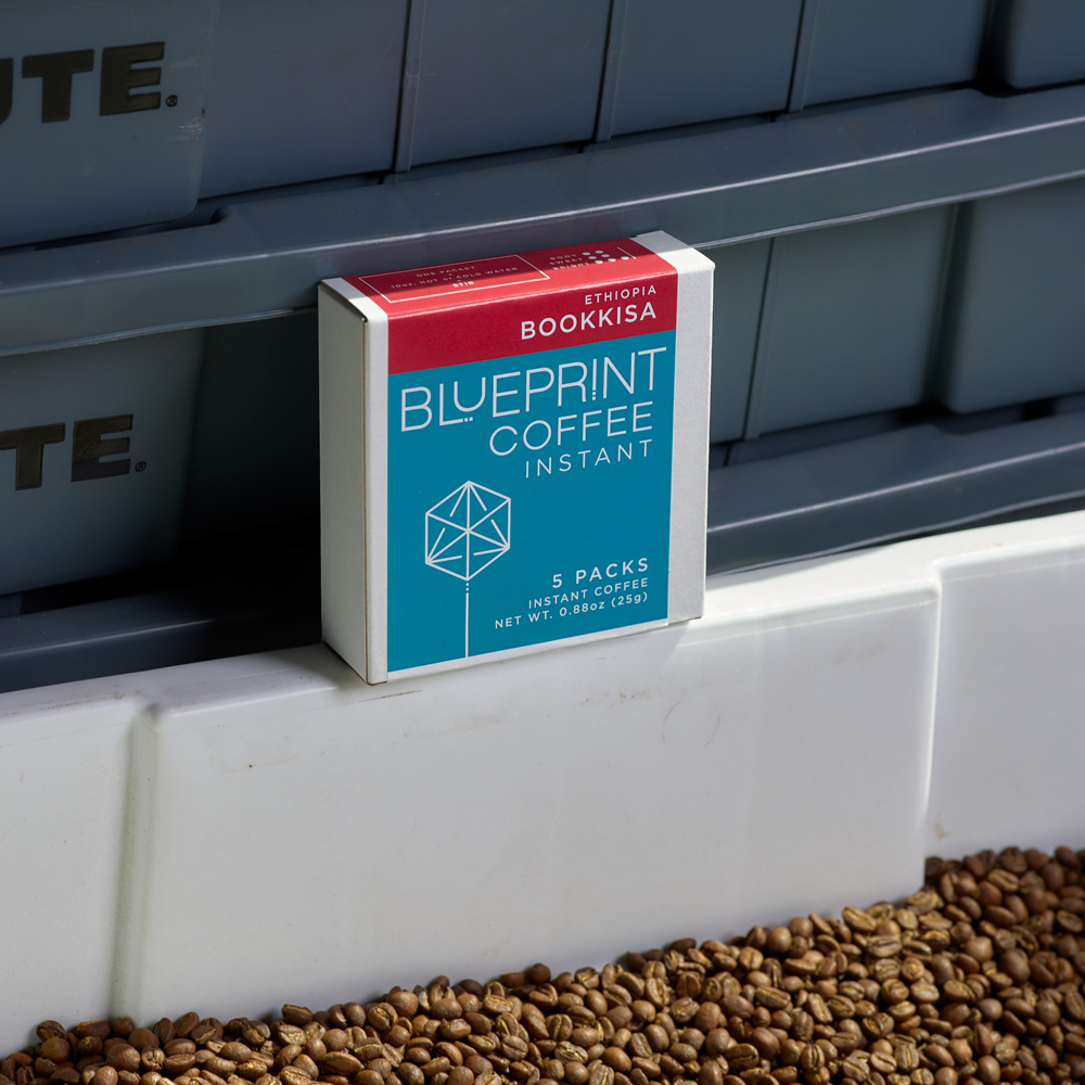 A box of Bookkisa, Ethiopia instant coffee rests on a tub of freshly roasted coffee beans.
