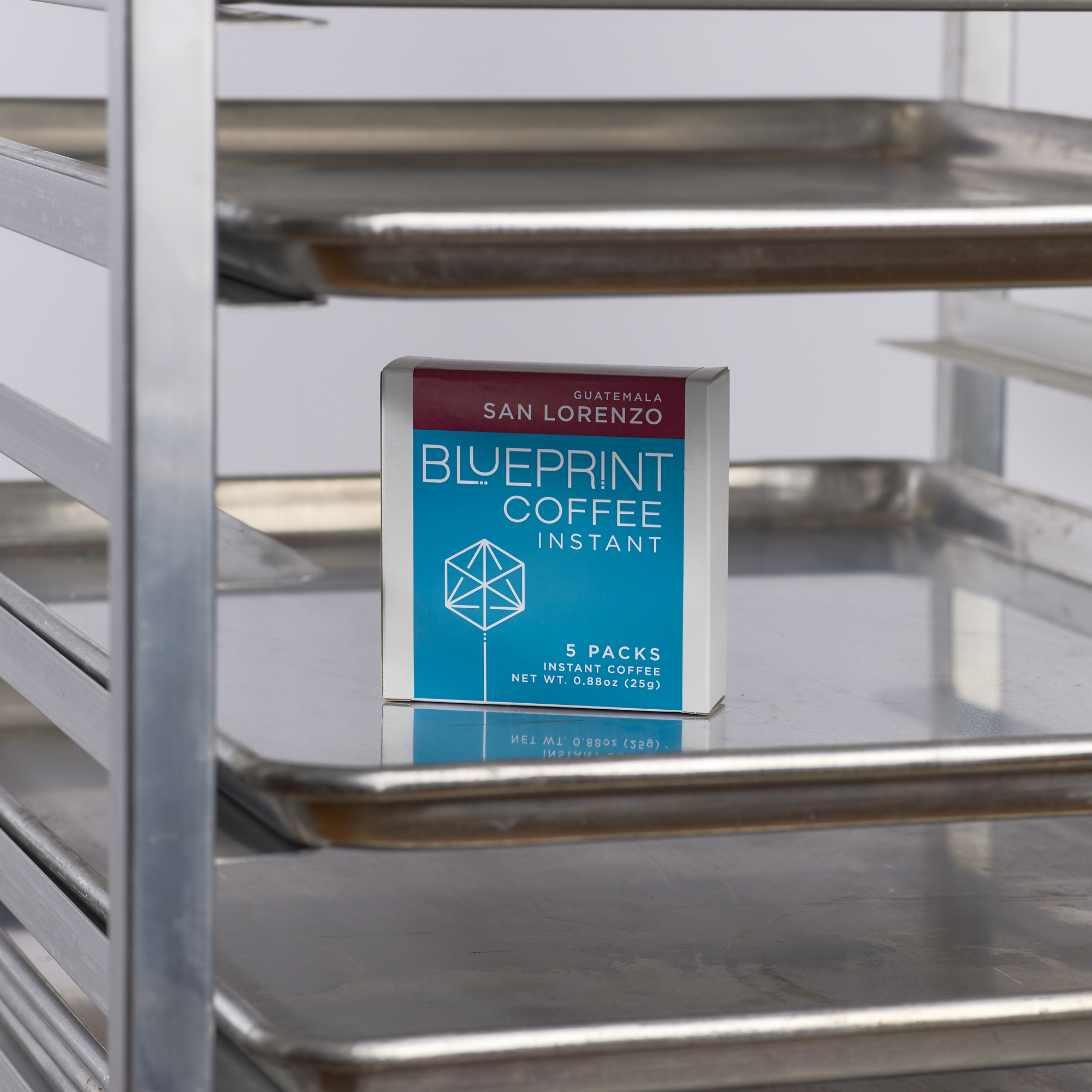 A white box containing San Lorenzo, Guatemala instant coffee roasted by Blueprint Coffee stands on a silver tray in a baker's speed rack. The box features the Blueprint Coffee logo in blue and the coffee name in red.