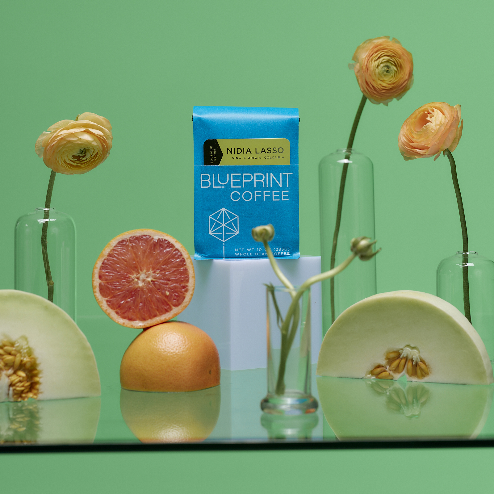 A blue bag of Nidia Lasso, Colombia roasted by Blueprint Coffee is placed in the center of a still life arrangement including orange flowers, cut grapefruit, and halved honeydew melons, with a bright green background