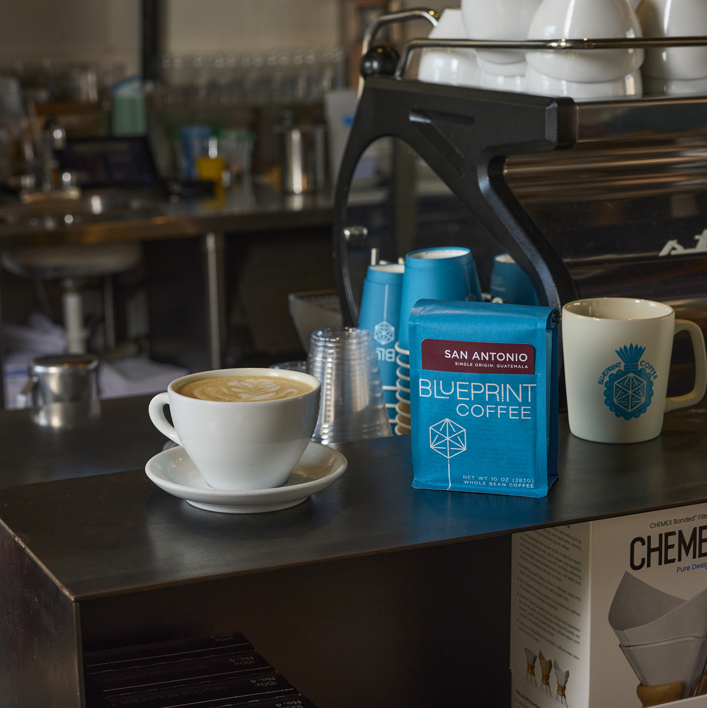 A blue bag of San Antonio, Guatemala by Blueprint Coffee sits on a dark bar in front of an espresso machine, beside a latte in a white ceramic mug